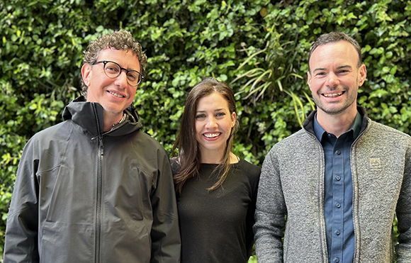 Three smiling adults standing together in front of a dense green leafy wall.