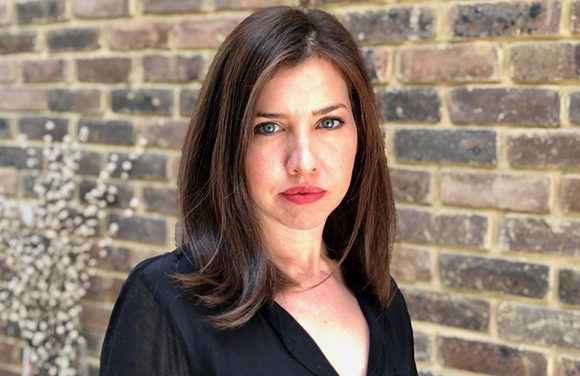 A woman with shoulder-length dark hair and blue eyes looking at the camera against a brick wall.
