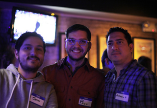 Three men smiling and wearing name tags in a dimly lit indoor setting.