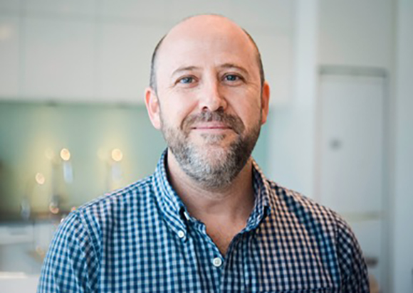 A man with a beard and short hair wearing a blue and white checkered button-down shirt.