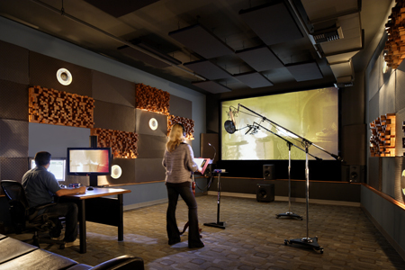 A musician stands in a recording studio facing a large projection screen and a microphone stand.