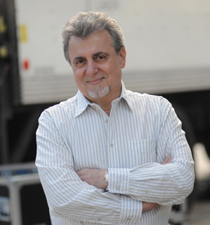 A middle-aged man with graying hair and a goatee wearing a light-colored striped shirt, smiling with arms crossed.