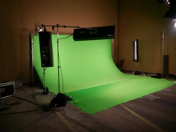 A large green screen backdrop curved on the floor with studio lights and equipment in a dark room.