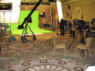 A film set featuring a large green screen, studio lights, a camera crane, and various production equipment.