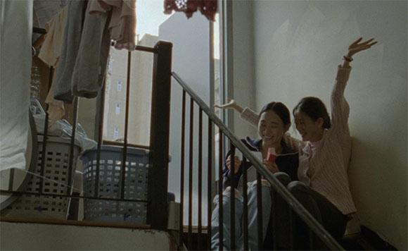 Two women sit on a staircase, laughing and gesturing with their hands raised.