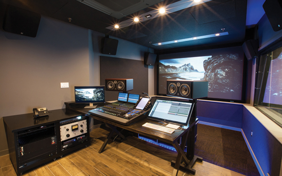A professional sound mixing studio with a large screen, mixing console, studio monitors, and rack equipment.