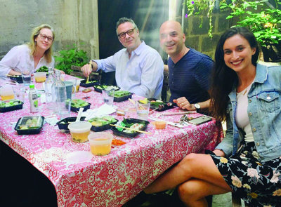 Four people smiling while sitting at a table covered in a pink patterned cloth with various food containers.