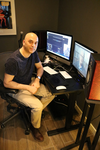 A man in a dark t-shirt sits at a desk with two monitors and studio speakers.