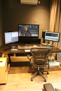 A desk with two computer monitors, a control panel, and an ergonomic office chair in a dimly lit room.