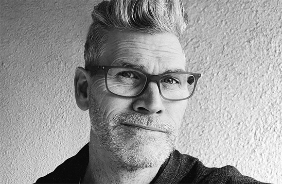 Black and white close-up portrait of a man with glasses, styled hair, and a short beard.