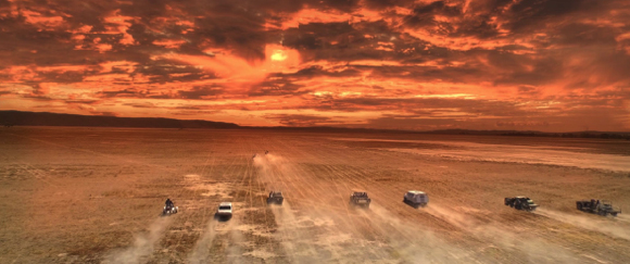Several vehicles drive across a dusty, flat landscape under a vibrant orange and red sunset sky.