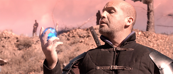 A bald man in dark armor holds a glowing blue sphere in his hand against a reddish, barren landscape.