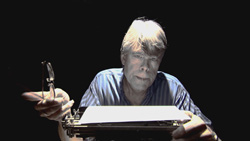 A man in a striped shirt holds glasses in one hand and rests the other on a typewriter against a black background.