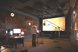 A person sits at a desk with monitors while another stands before a large screen and microphone in a studio.