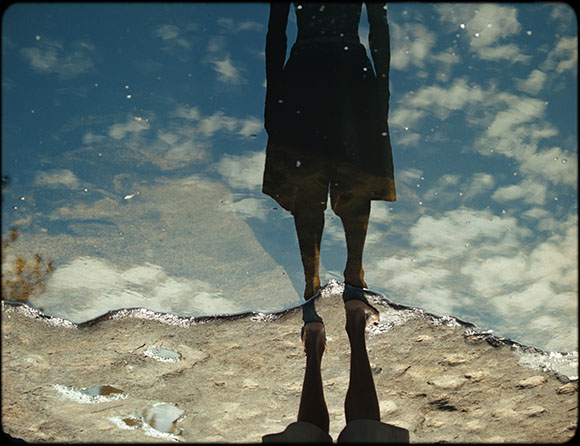 Silhouette of a person reflected in a puddle under a blue sky with white clouds.