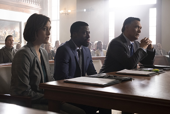 Three people in professional attire sit at a long wooden table in a courtroom.