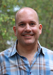A smiling man with a shaved head wearing a blue and tan plaid button-down shirt.