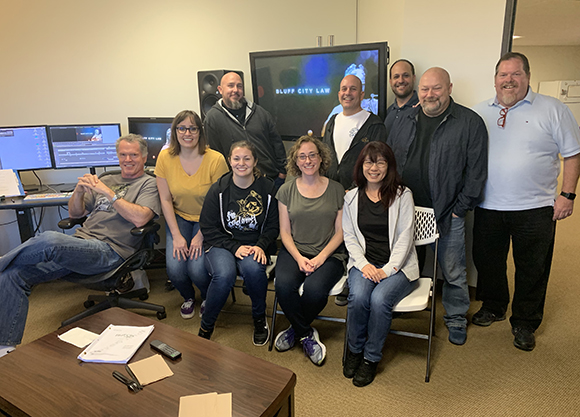 A group of ten diverse adults posing together in an office with computer monitors and a television in the background.
