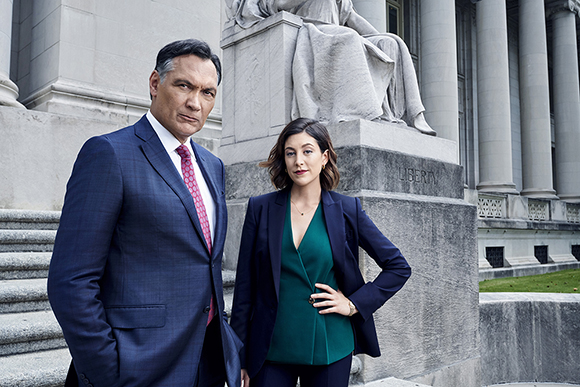 A man in a blue suit and a woman in a dark blazer and green top pose before a stone building and statue.