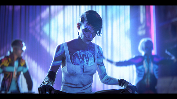 A person with glowing blue tattoos and facial implants sits in a dark room with neon lighting.