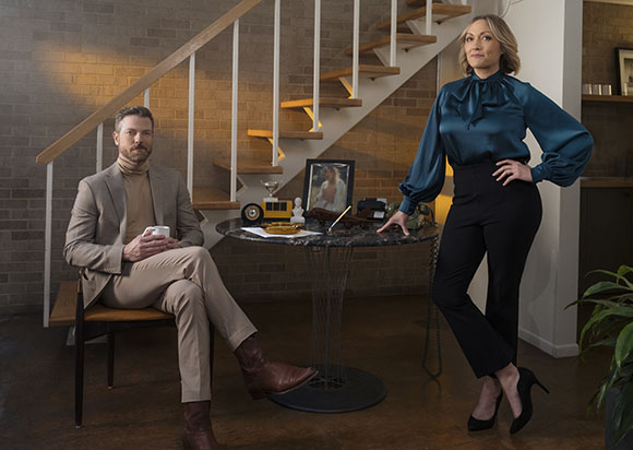 A man in a tan suit sits at a table while a woman in a teal blouse and black pants stands beside him.
