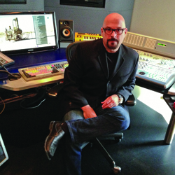 A man with glasses and a goatee sitting in a music studio with mixing consoles and monitors.
