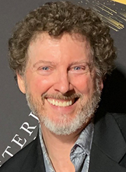 A smiling man with curly brown hair and a graying beard wearing a dark blazer and patterned shirt.