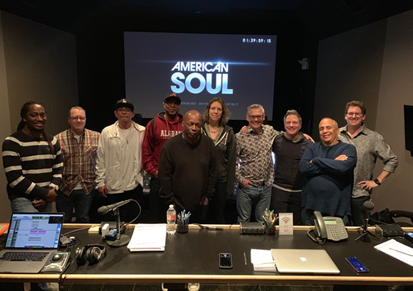 A group of ten diverse people standing together in a dark room in front of a screen titled American Soul.