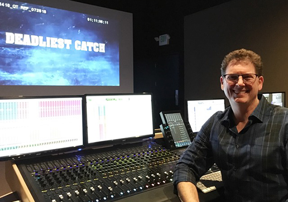 A man in a plaid shirt smiles next to a large audio mixing console and multiple computer monitors.