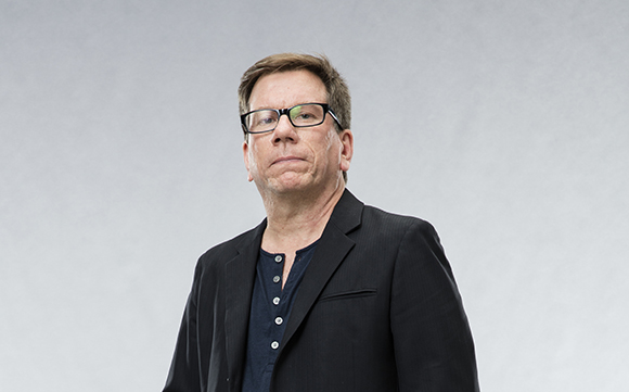 A man with glasses wearing a black blazer and a dark button-down shirt against a gray background.