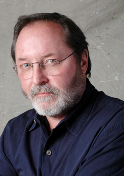 A man with short brown hair, glasses, and a graying beard wearing a dark blue button-down shirt.