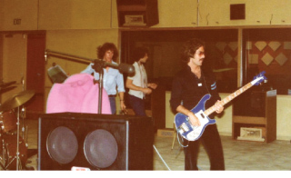 A man plays a blue electric bass guitar in a music studio with other musicians in the background.