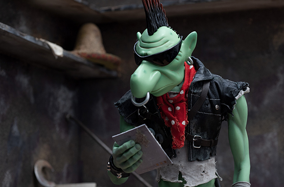 A green goblin figure with a mohawk, sunglasses, and a red bandana wearing a black vest.