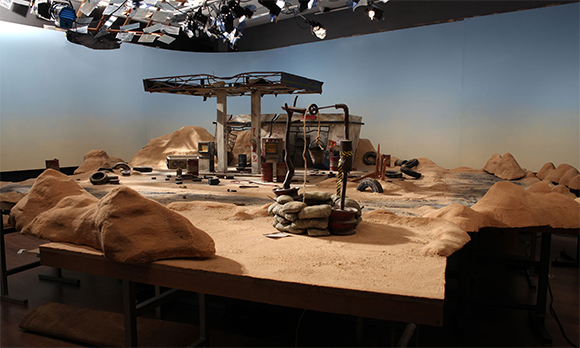 A miniature desert diorama with a ruined gas station, sand dunes, and scattered debris on a large table.