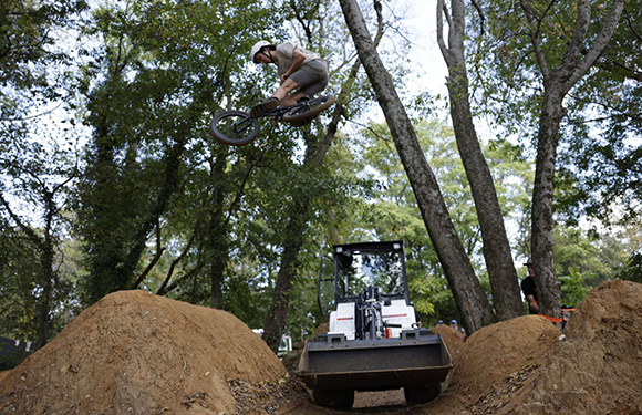 A person on a bicycle jumps high in the air above dirt mounds and a small loader in a wooded area.
