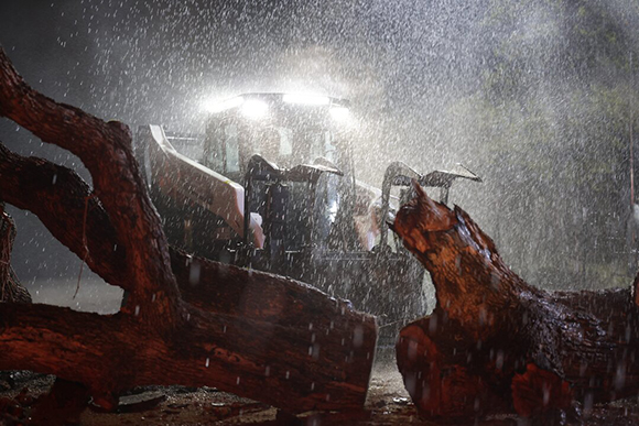 A heavy machine with bright headlights operates in a heavy downpour amidst large fallen logs.