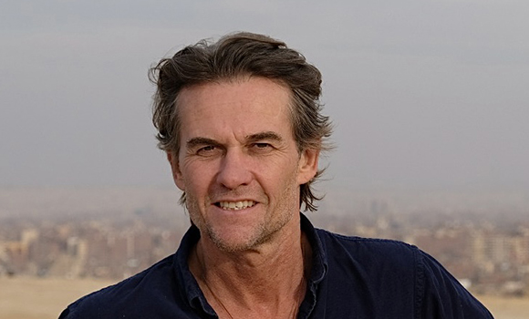 A man with wavy brown hair smiling outdoors against a blurred cityscape background.