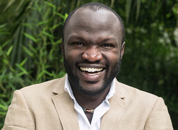 A smiling Black man with a beard wearing a tan blazer and white shirt against a green leafy background.