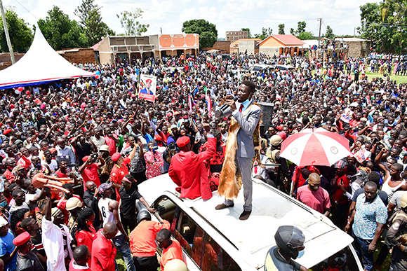A man in a grey suit stands on a white car roof addressing a massive outdoor crowd.