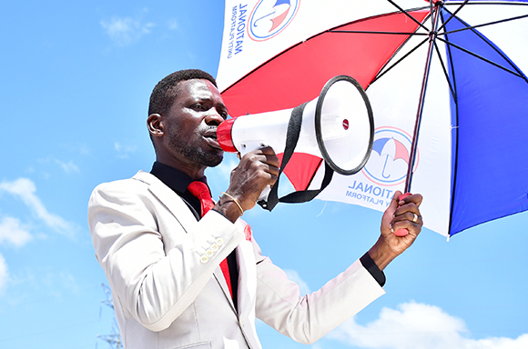 A man in a white suit speaks into a megaphone while holding a red, white, and blue umbrella.