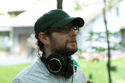 A man with a beard, glasses, and a green cap wearing headphones around his neck.