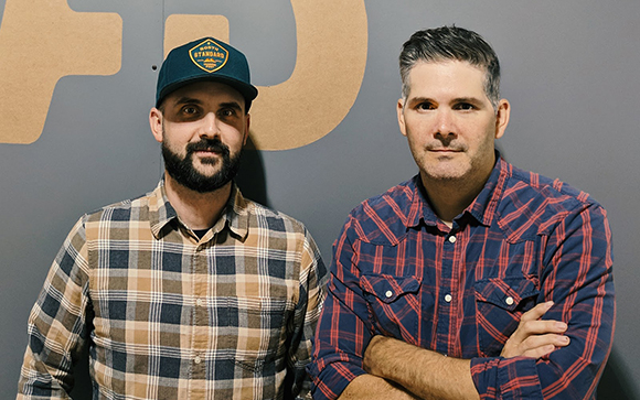 Two men posing together, one in a plaid shirt and cap, the other in a red plaid shirt with arms crossed.