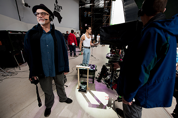 A man in a black hat and blue jacket stands on a film set near a camera operator and equipment.