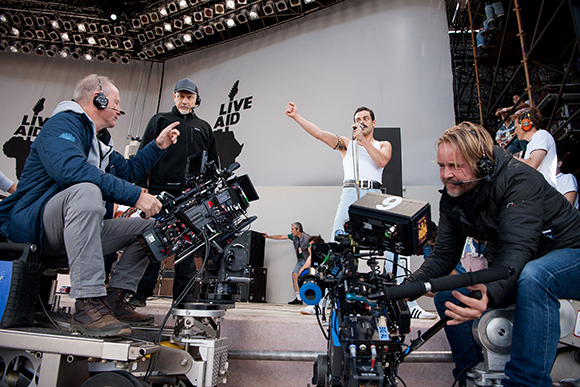 Camera operators film a male singer performing on a stage with Live Aid branding in the background.