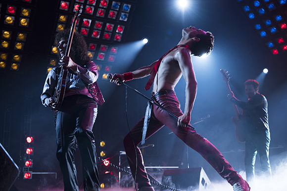 A shirtless male singer leans back holding a microphone while two musicians play instruments on a lit stage.
