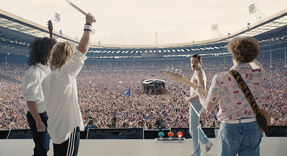 Four musicians on a stage perform for a massive, dense crowd in a large outdoor stadium.