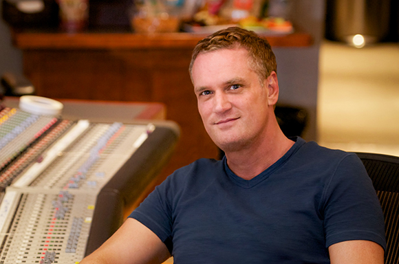 A man in a blue v-neck shirt sits in a studio next to a large mixing board.