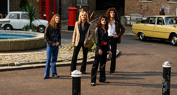 Four young men with long hair posing in a street with vintage cars and a red telephone box.