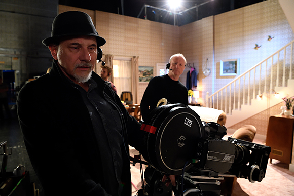 A man in a black hat operates a large film camera on a movie set with a staircase in the background.