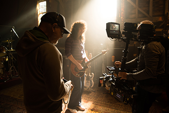 A film crew uses a professional camera to film a woman playing an electric guitar under bright studio lighting.